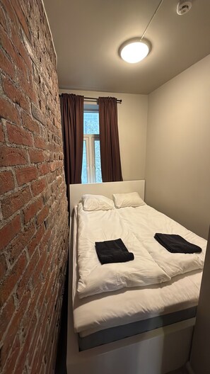 A bedroom with a brick wall, a window with curtains, a bed with white bedding, and a ceiling light. - Super central apartment in Trondheim, steps from city centre & restaurants! (Trøndelag)