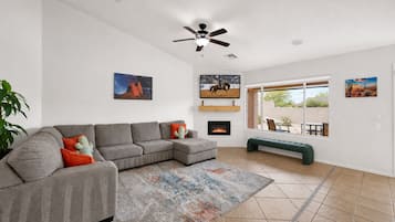 Ein Wohnzimmer mit einem grauen Ecksofa, einem Kamin, einem Deckenventilator und einem großen Fenster mit Blick auf eine Wüstenlandschaft.