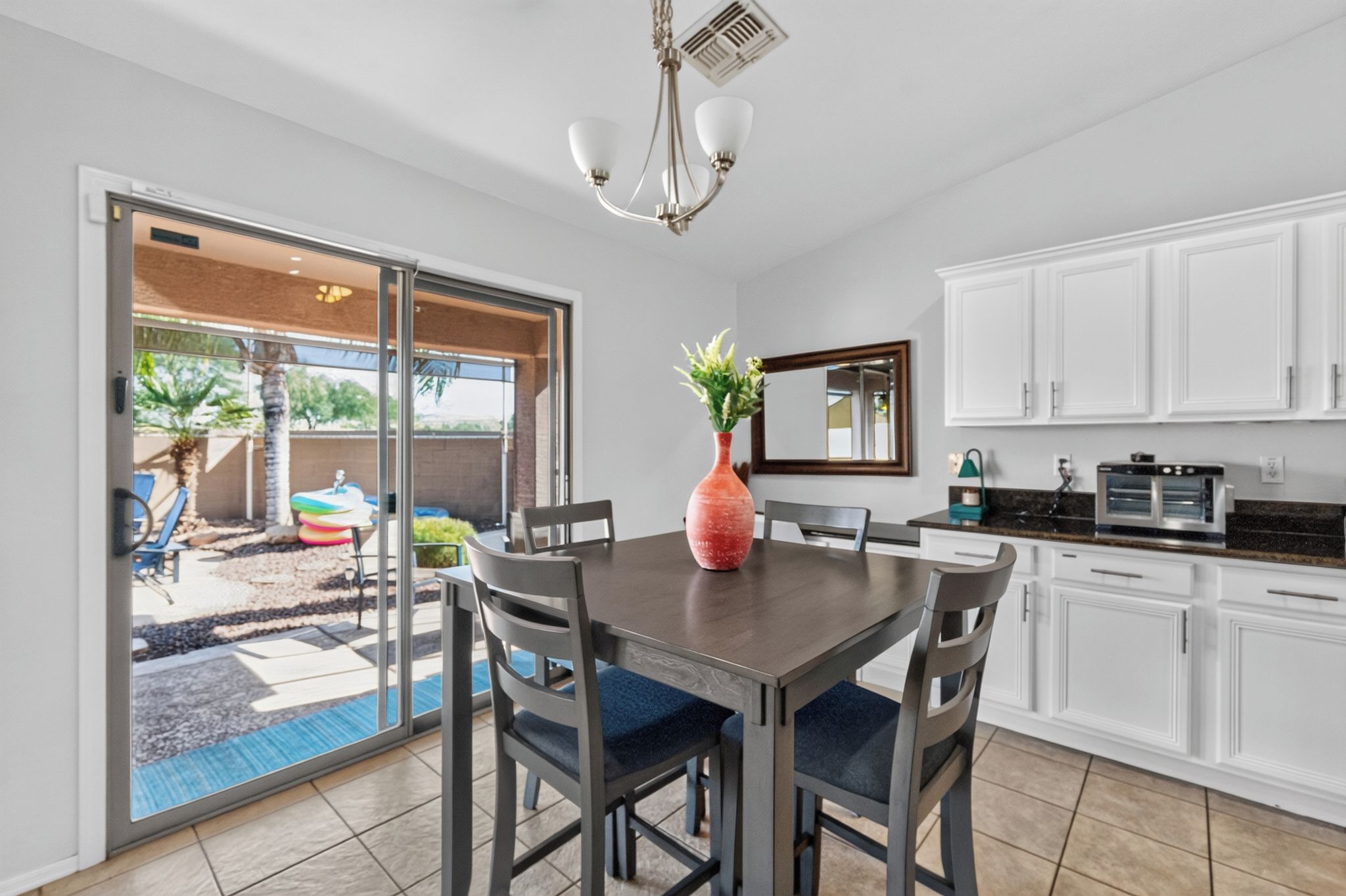 A modern kitchen with white cabinets, a dining table with chairs, a red vase, and a sliding glass door leading to an outdoor area.