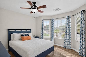 Un dormitorio con una cama, una mesita de noche, un ventilador de techo y cortinas estampadas.