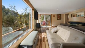A spacious living room with a large window, a sofa, a bench, and a television. - The Salmon Hole (Washougal)