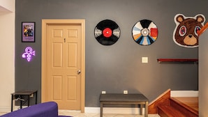 Ett rum med en dörr av trä, en bänk, en lila soffa och väggdekorationer som inkluderar en vinylskiva, en nallebjörn och en neonskylt.