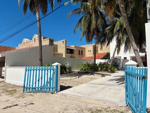 A gated entrance with a blue fence, palm trees, and buildings with a beige facade and red-tiled roofs.