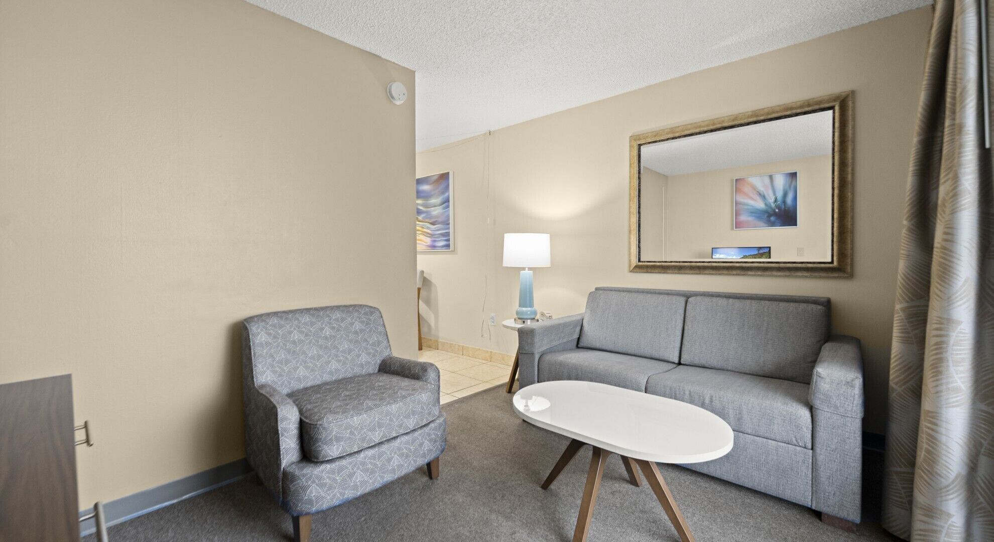 A hotel room with a grey armchair, a grey sofa, a white coffee table, and a lamp on a side table.