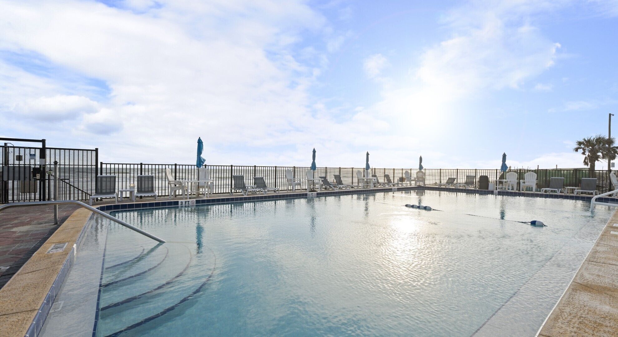 Ein Schwimmbecken mit Liegestühlen und Sonnenschirmen, umzäunt, mit Blick auf das Meer und einer Statue im Hintergrund.