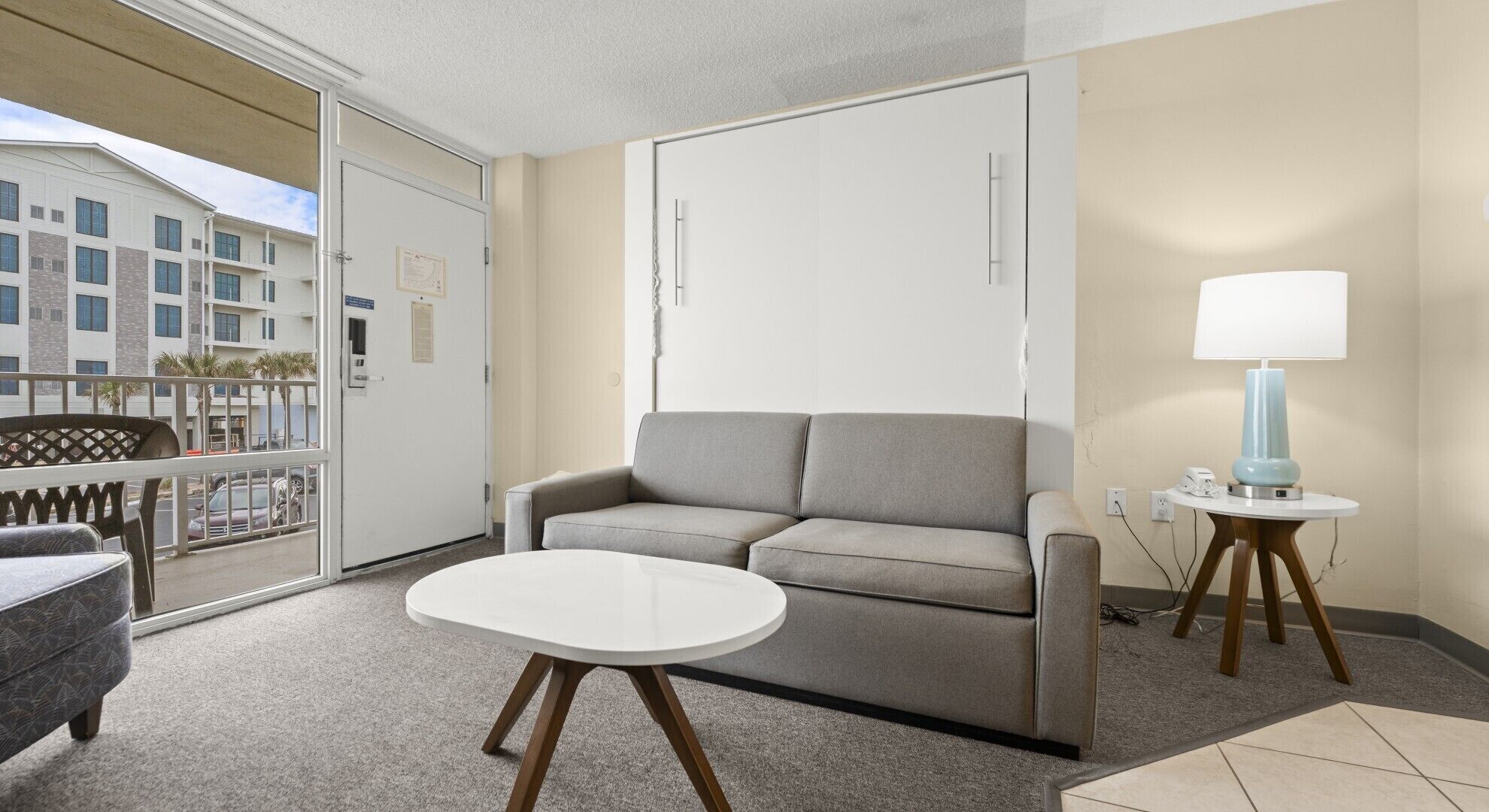 Une chambre d’hôtel moderne avec un canapé gris, une petite table ronde et une lampe. Elle dispose d’un balcon avec vue sur des immeubles et une voiture.