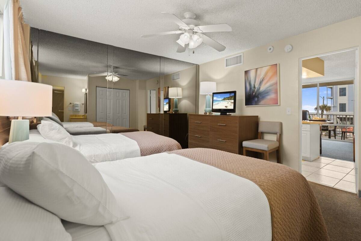 A hotel room with two beds, a ceiling fan, a TV, and a view of the outside balcony.