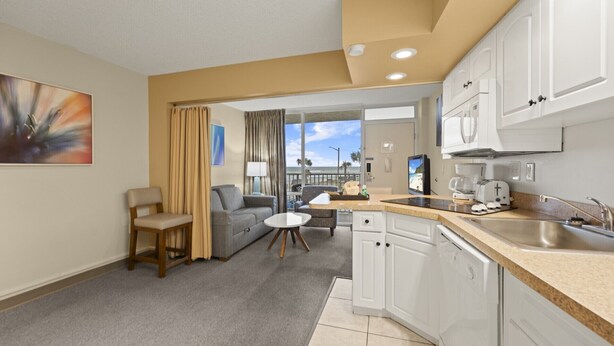 Una habitación de hotel moderna con kitchenette, una zona de estar con sofá y sillas, un balcón con vistas y un cuadro en la pared.