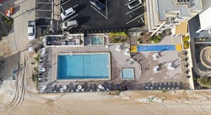 An aerial view of a hotel with a large rectangular pool, multiple lounge chairs, and a parking area.