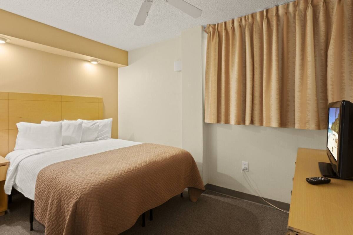 A hotel room with a bed, a bedside table with a lamp, a TV on a stand, and a window with curtains.
