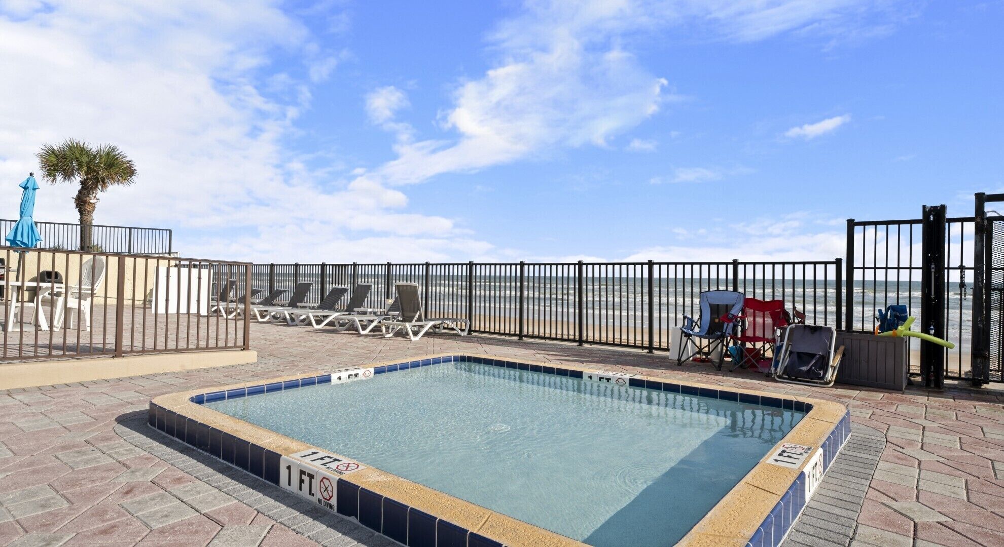 A swimming pool with a 1 ft depth limit, surrounded by lounge chairs and a fence overlooking the beach.