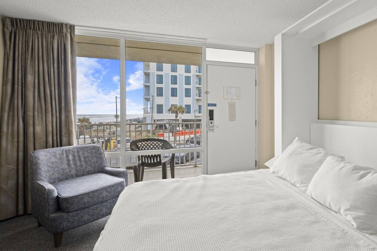 A hotel room with a bed, a chair, and a view of a building and beach.