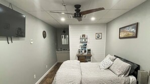 A bedroom with a bed, a desk, a chair, a ceiling fan, a TV, and a bathroom in the background.