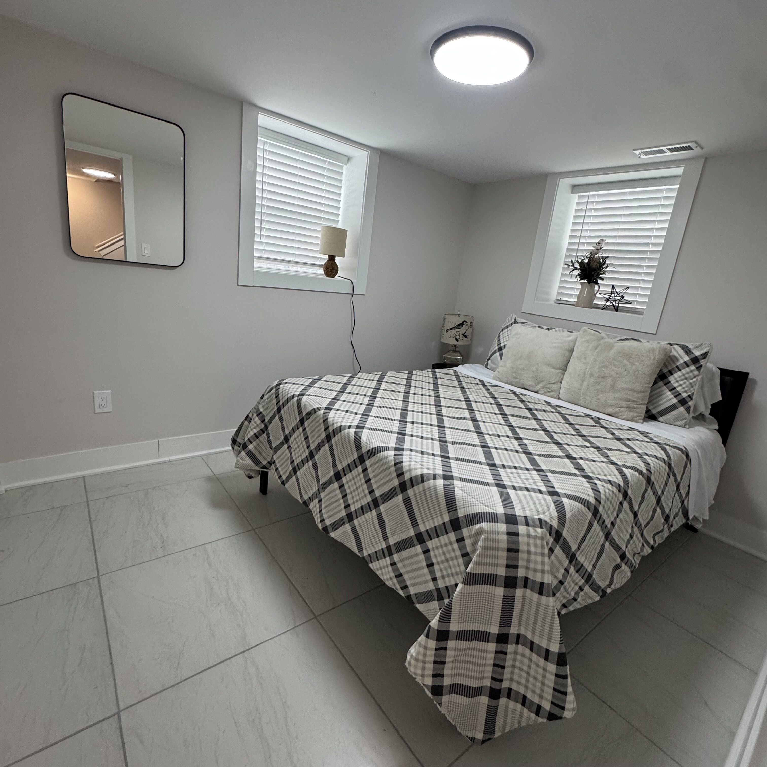 A bedroom with a bed, two windows with blinds, a bedside table with a lamp, a wall-mounted mirror, and a ceiling light.