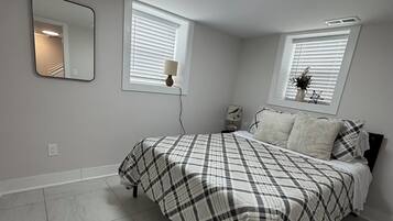A bedroom with a bed, two windows with blinds, a bedside table with a lamp, a wall-mounted mirror, and a ceiling light.