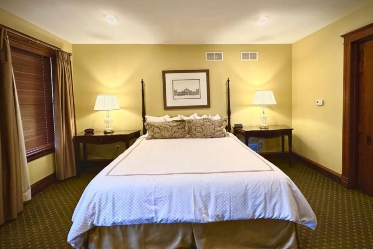 Une chambre d’hôtel avec un grand lit, deux tables de chevet, deux lampes, un tableau encadré au mur et une fenêtre avec des rideaux.