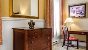Une chambre d’hôtel avec une commode en bois, une chaise, un bureau, une lampe, un tableau encadré et une fenêtre avec des rideaux.