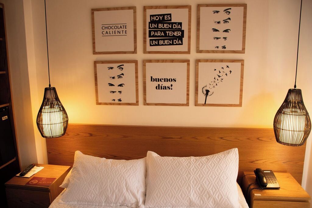 A hotel room with a wooden headboard, a bed with white pillows, two bedside tables with lamps, and motivational wall art.