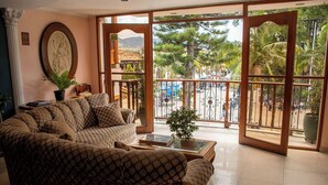 Una sala de estar con un sofá estampado, una mesa de centro y vistas a un balcón con mobiliario exterior y palmeras.