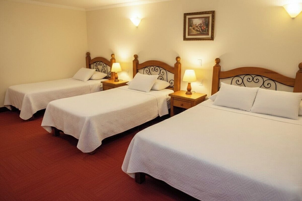 A hotel room with three single beds, each with a wooden headboard and a bedside table with a lamp.