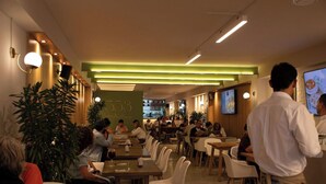 Interior de restaurante moderno con varias mesas y sillas, una pared de acento verde y una pantalla de televisión mostrando comida.