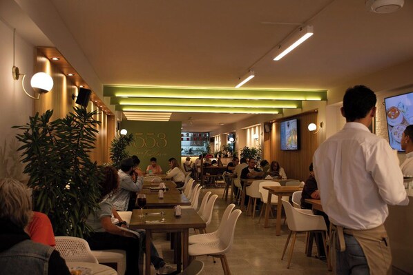 Interior de restaurante moderno con varias mesas y sillas, una pared de acento verde y una pantalla de televisión mostrando comida.