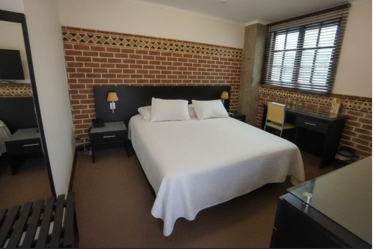 A hotel room with a brick-patterned wall, a bed with white linens, a nightstand with a lamp, and a window with blinds.