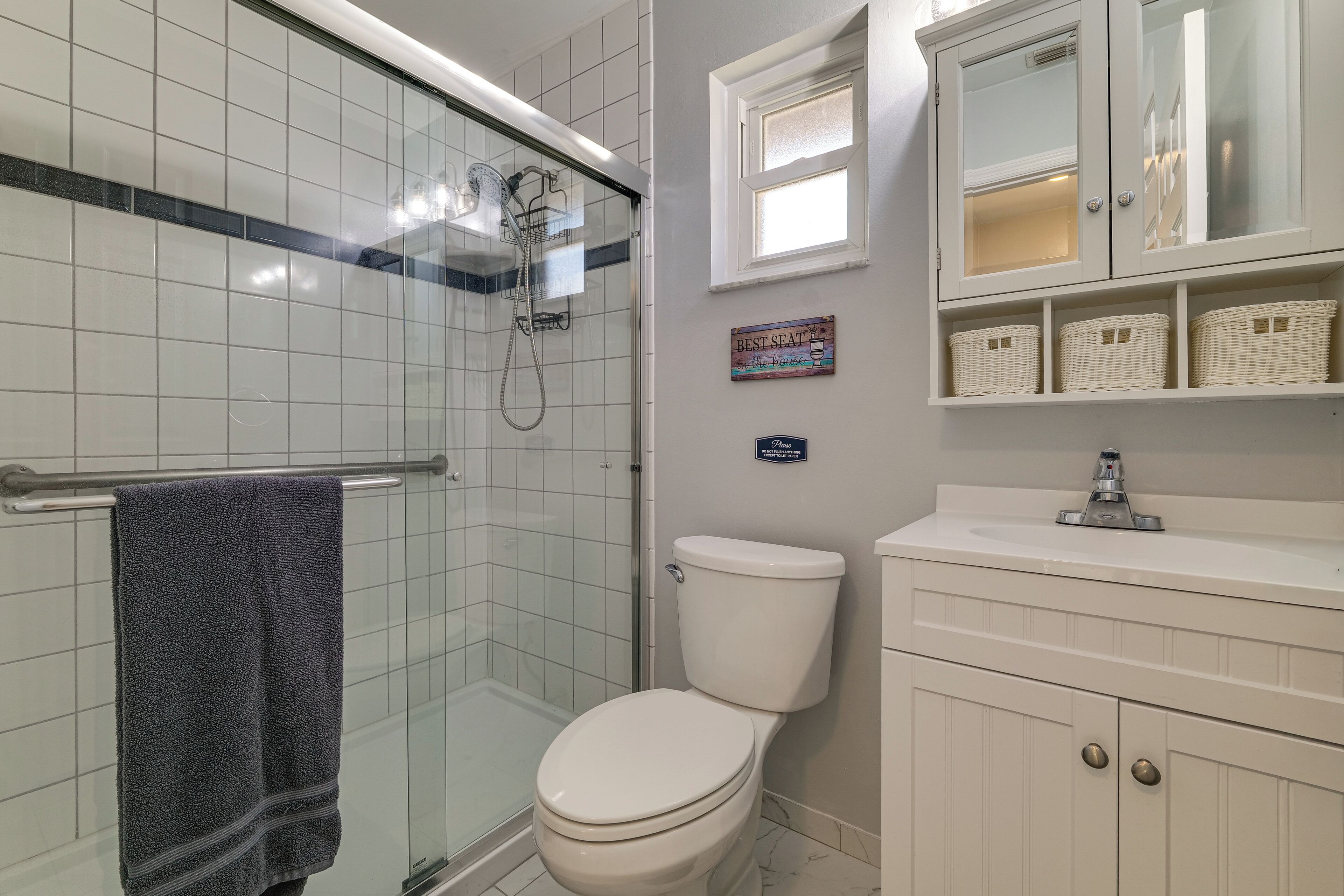 Ein Badezimmer mit Duschkabine aus Glas, einer weißen Toilette, einem Waschtisch und Körben aus Weidengeflecht auf Regalen.