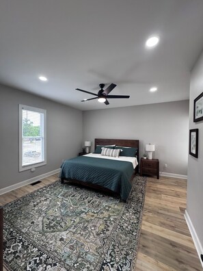 Une chambre à coucher comprenant un lit, un ventilateur de plafond, une table de chevet, une lampe, une fenêtre avec des stores et un tapis à motifs.