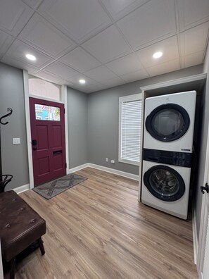 A laundry room with a front-loading washer and dryer, a red door, and a window with blinds. - Retreat on Bridge Street - 3 Bed, 2 Bath Luxury Apartment (Tunkhannock)