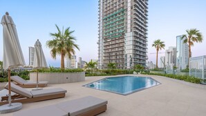 A rooftop pool area with lounge chairs and umbrellas, surrounded by palm trees and modern buildings. - Stayshort - Modern 1 Bedroom With Private Jacuzzi in JVC That Sleeps 2 (Dubai)