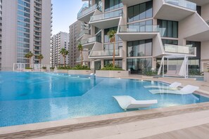 En moderne swimmingpool med liggestole, omgivet af høje bygninger med altaner og palmer.