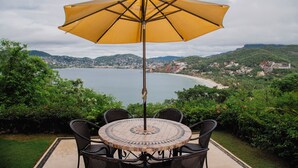 A round table with four chairs and a yellow umbrella, overlooking a coastal town with a beach and hills. - Zihua Vista Bay Principal: Stunning La Ropa Views, Infinity Pool & Gourmet Kitch (Zihuatanejo)