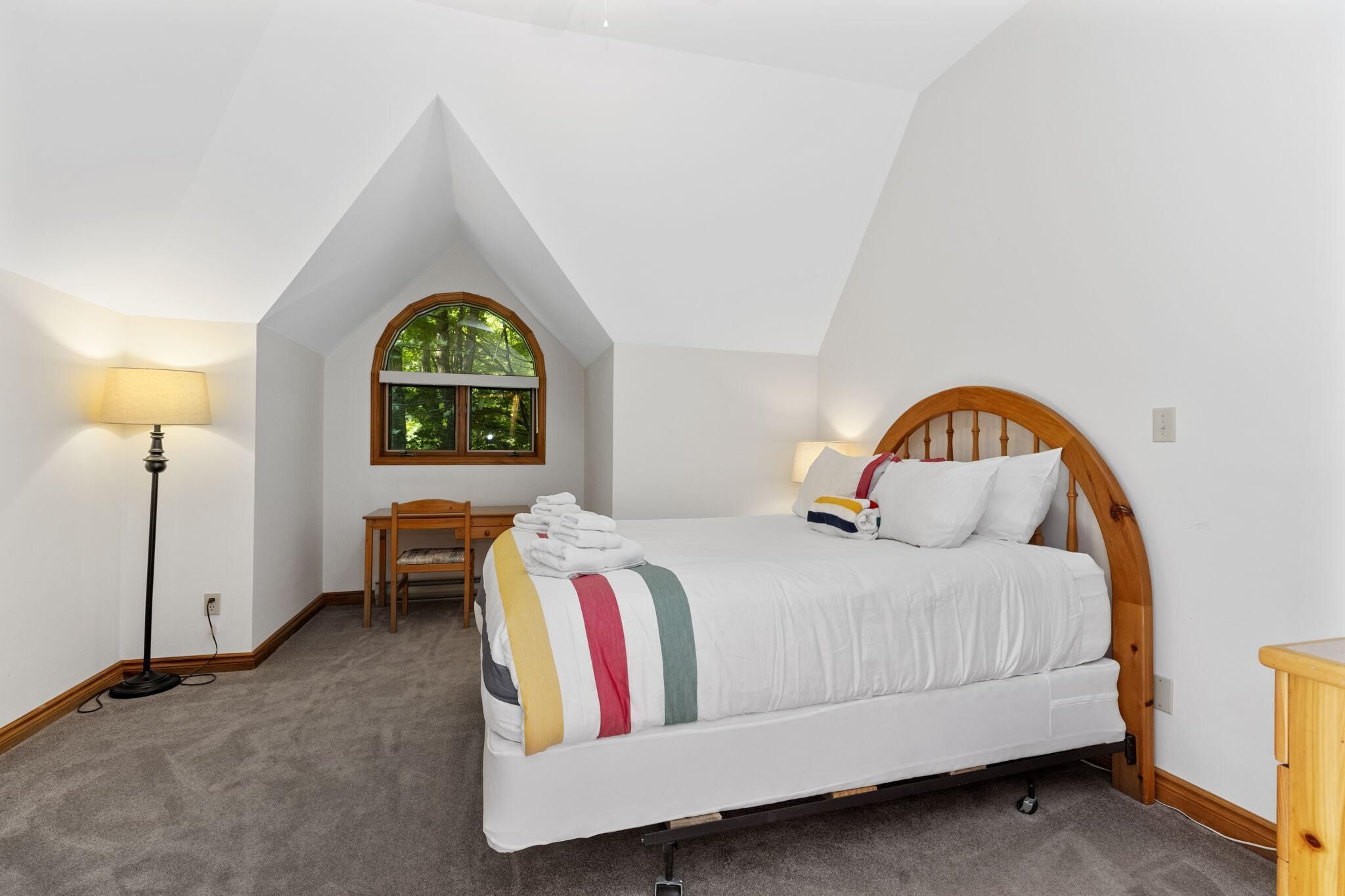 Un dormitorio con cama, escritorio y silla. Se ve una ventana con vista a árboles.