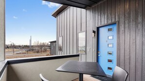 Un balcon avec une table et des chaises, une porte bleue avec des panneaux numérotés, et une vue sur une route et des bâtiments en arrière-plan.