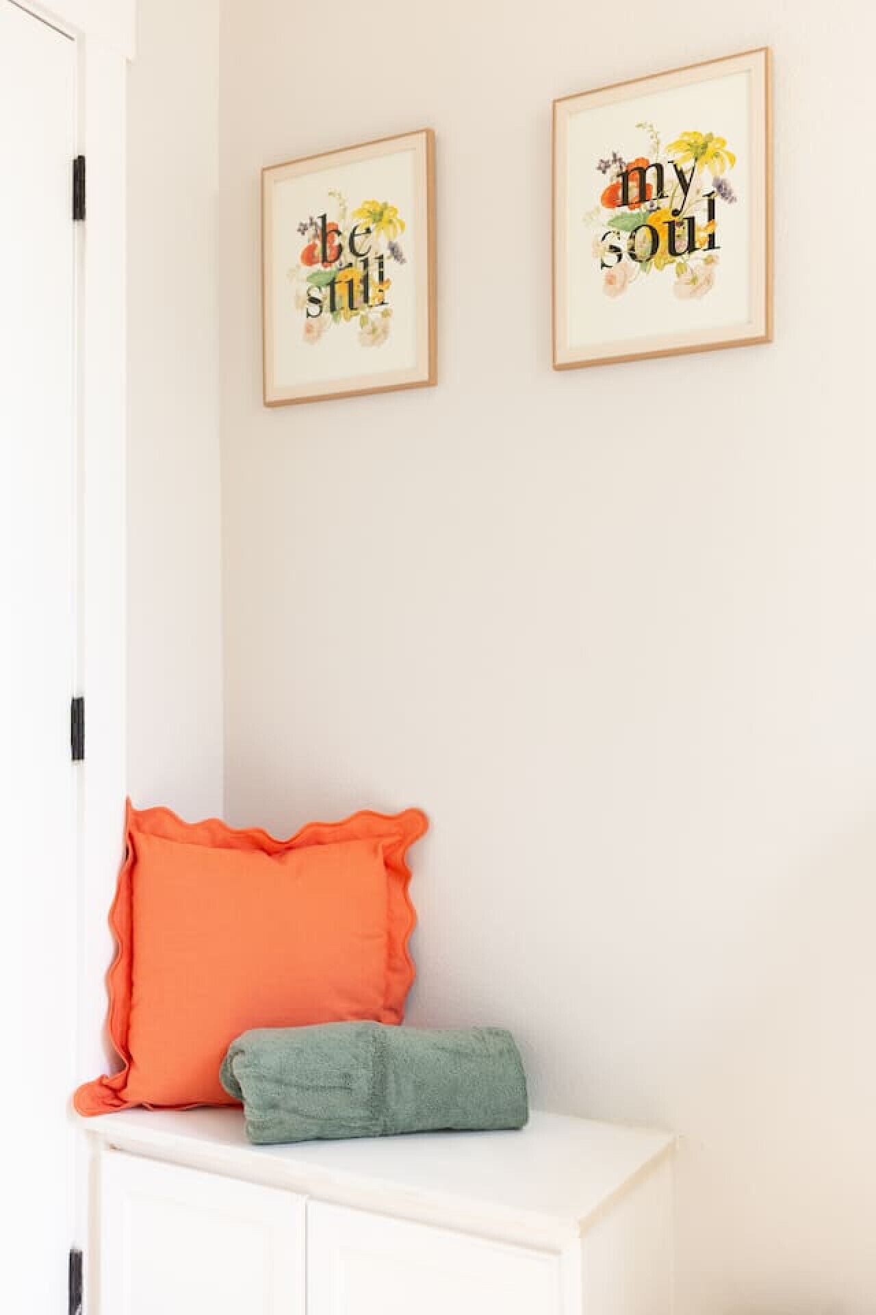 Una habitación con un armario blanco, un cojín naranja y un cojín verde encima. Dos obras de arte enmarcadas con motivos florales y texto están colgadas en la pared.