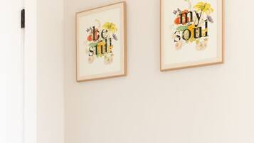 Una habitación con un armario blanco, un cojín naranja y un cojín verde encima. Dos obras de arte enmarcadas con motivos florales y texto están colgadas en la pared.