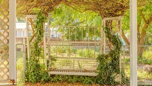 Eine hölzerne Bank unter einer mit Wein bewachsenen Pergola, umgeben von einem Ziegelweg und einem Zaun.