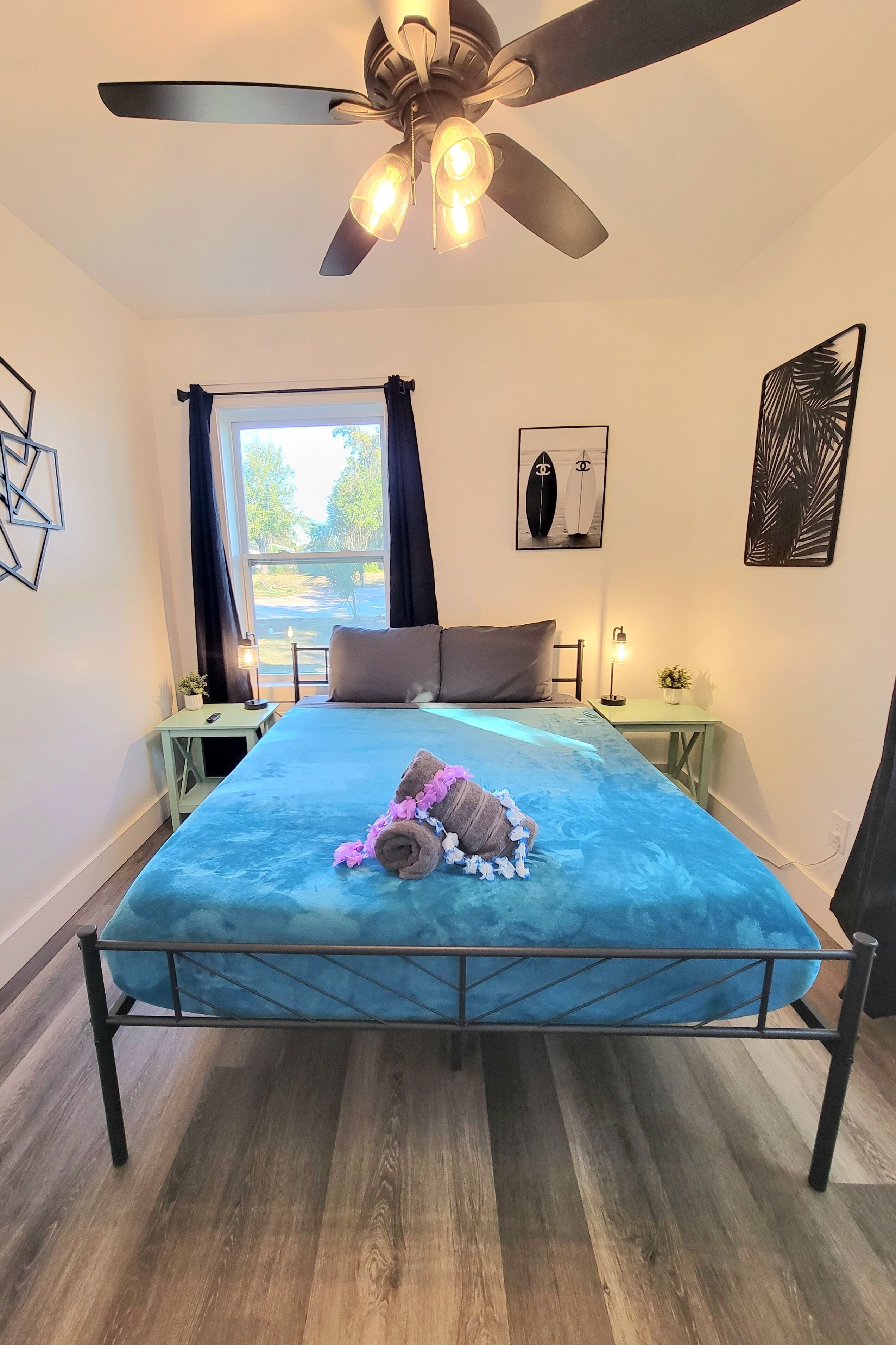 Une chambre à coucher avec un lit, un ventilateur de plafond, une fenêtre avec des rideaux, une table de chevet avec une lampe et un mur orné d’un tableau encadré.