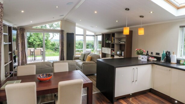 A modern kitchen with a central island, dining area with a table and chairs, and a view of the outdoors through large windows. - Plas Marchog Lodge (Pentraeth)