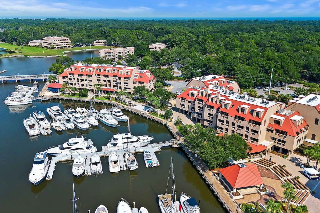 A marina with multiple boats docked, surrounded by buildings with red roofs and a lush green landscape in the background.