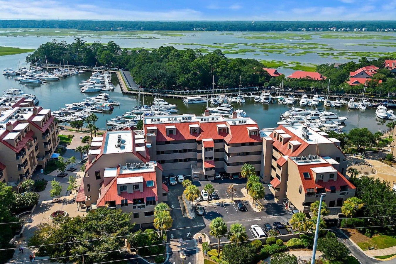 A marina with multiple boats docked, surrounded by buildings with red roofs and a parking area.