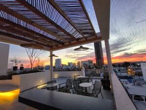 A rooftop terrace with outdoor seating, a view of the city skyline, and a sunset.
