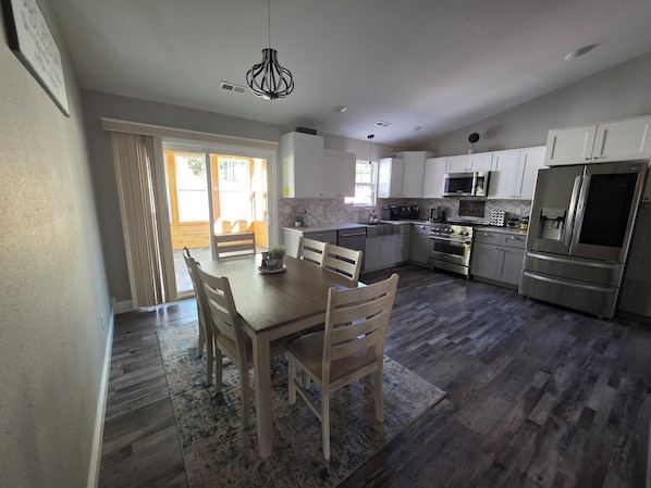 A modern kitchen with white cabinets, stainless steel appliances, and a wooden dining table with chairs. - The Gathering Place at Ft. Wood (Waynesville)