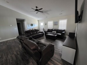 A spacious living room with a sectional sofa, a recliner, and a flat-screen TV mounted on the wall. - The Gathering Place at Ft. Wood (Waynesville)