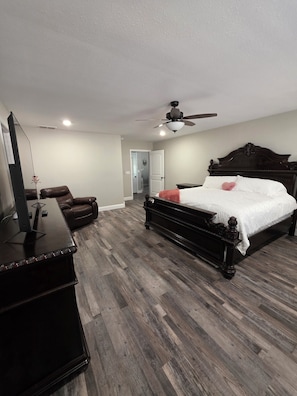 A bedroom with a bed, a dresser, a TV, a ceiling fan, and a door leading to another room. - The Gathering Place at Ft. Wood (Waynesville)