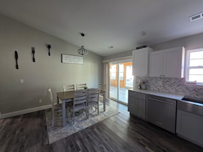 A modern kitchen with a dining area, featuring a wooden table and chairs, a chandelier, and a herringbone tile backsplash. - The Gathering Place at Ft. Wood (Waynesville)