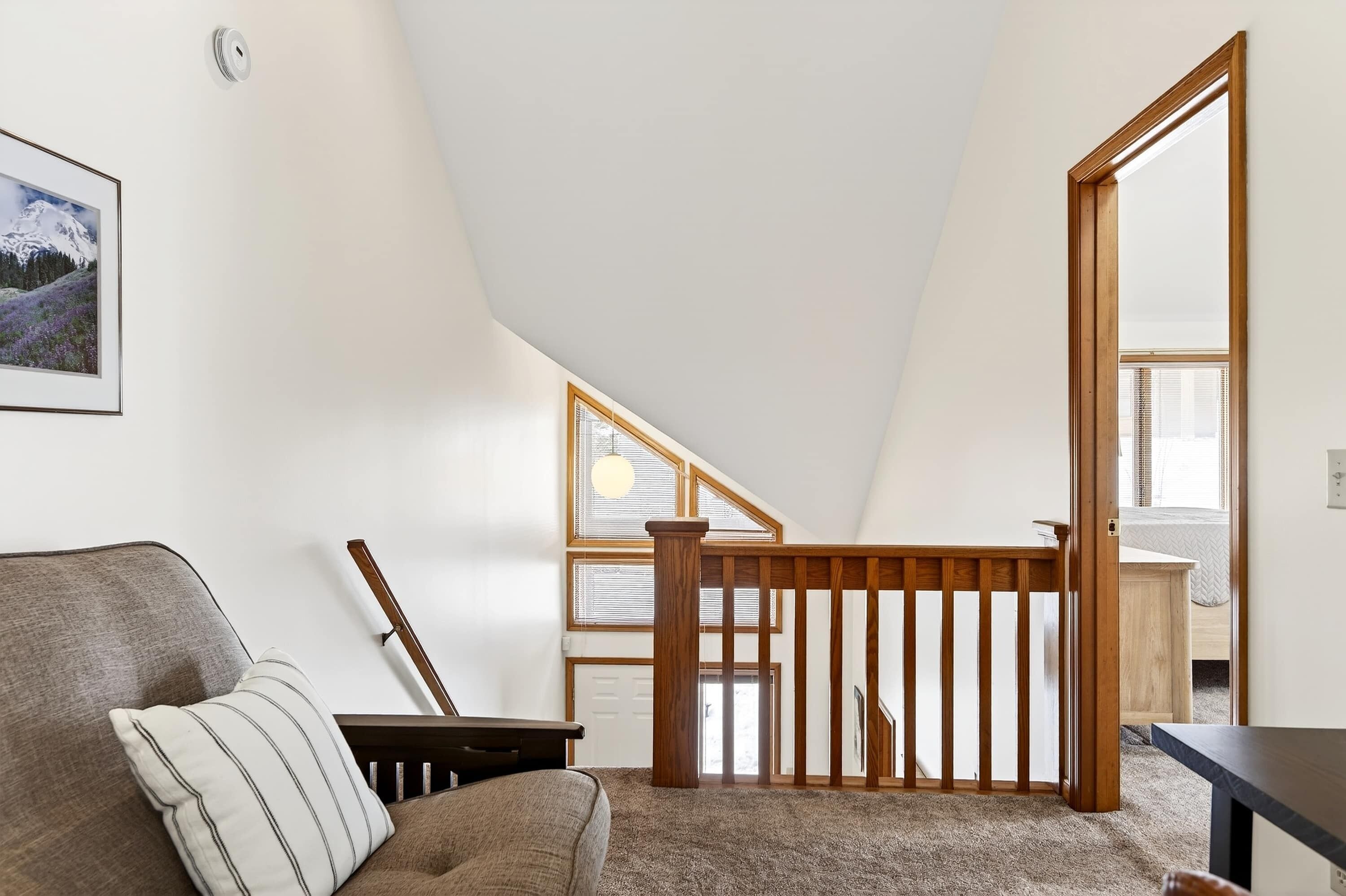 A room with a sloped ceiling, a wooden staircase, a cushioned chair, a small table, and a framed picture on the wall.
