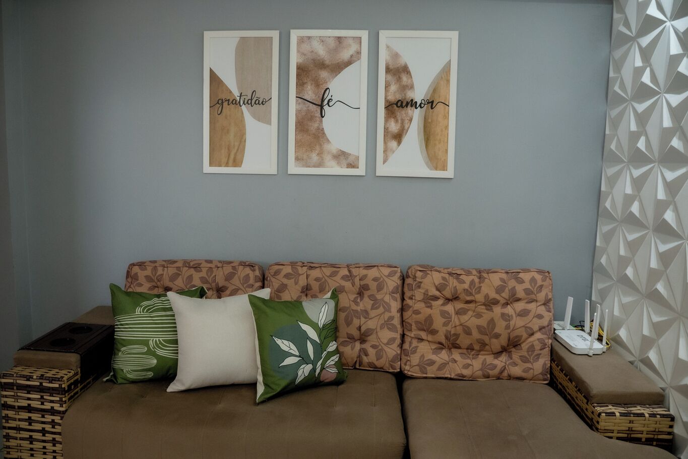 A living room with a brown sofa, three framed artworks with Portuguese words, and a geometric patterned wall.