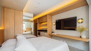 Habitación de hotel moderna con una cama grande, un televisor de pantalla plana en la pared, una pequeña cocina y un baño visible a través de una puerta abierta.
